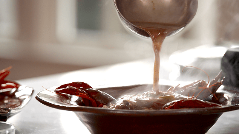 A steaming brown sauce is poured over crawfish in a brown bowl, creating a warm and appetizing scene. Sunlight softly illuminates the dish.