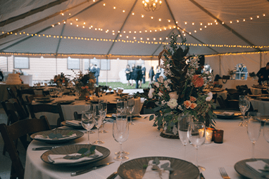 A beautifully set wedding reception under a white tent with hanging string lights. Tables are adorned with elegant floral centerpieces and neatly arranged tableware. Soft lighting creates a warm and festive atmosphere.