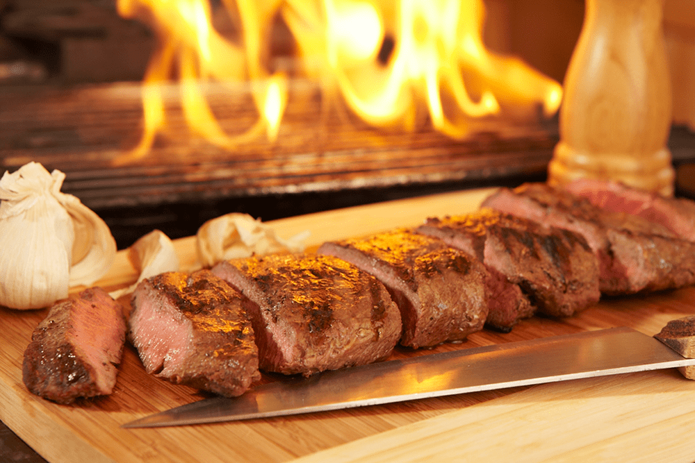 Sliced grilled steak on a wooden board with garlic bulbs beside it, a knife in the foreground, and flames from a grill in the background.
