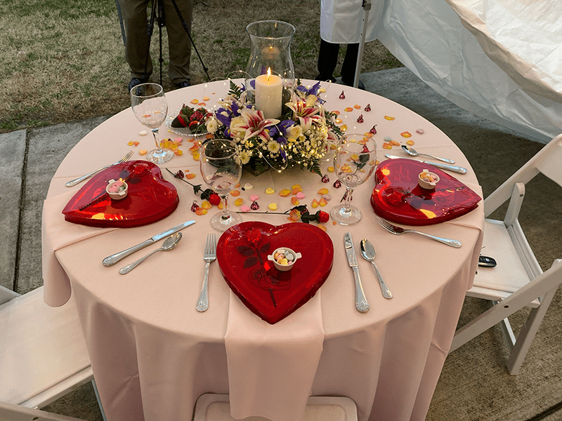 A round table with a pink tablecloth, set for two. Heart-shaped red trays hold small treats. A floral centerpiece with a candle adds elegance, surrounded by scattered petals, creating a romantic atmosphere.